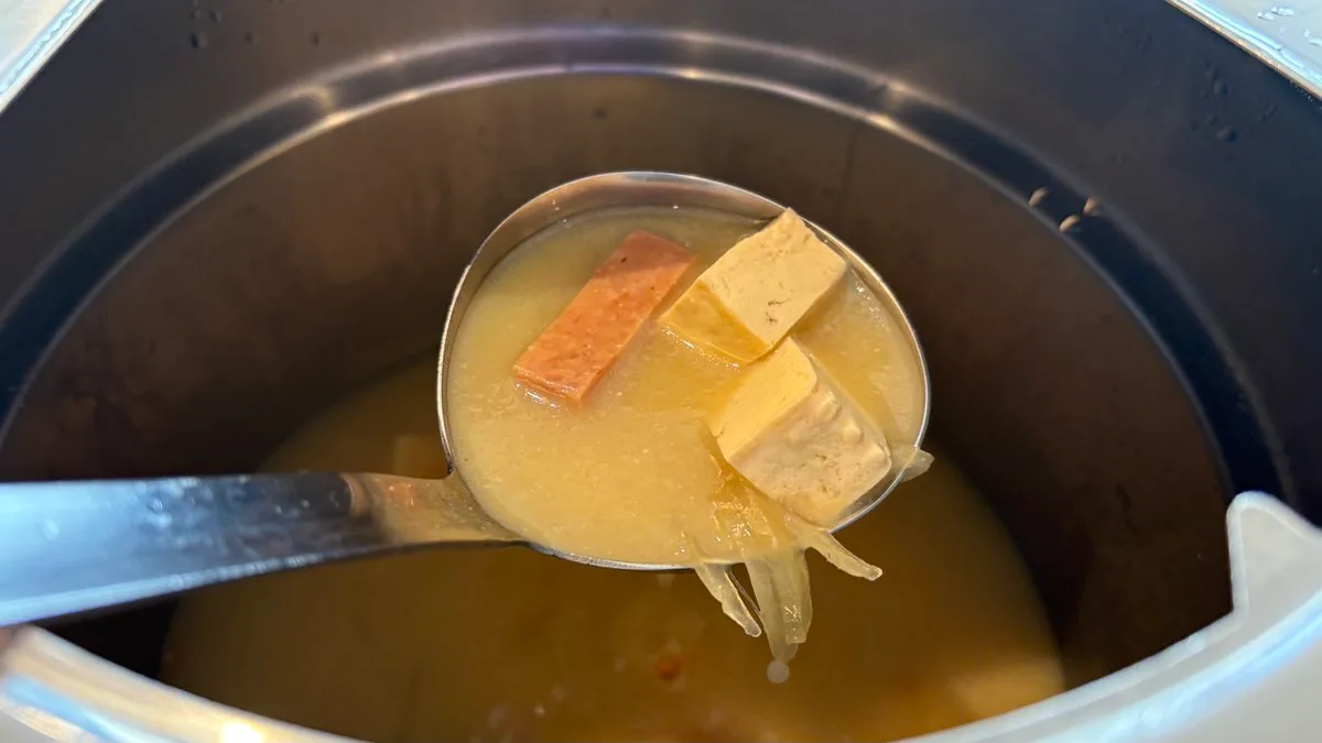 A ladle lifted from a pot of Okinawan miso soup holding a slice of spam, cubes of island tofu and strands of onion