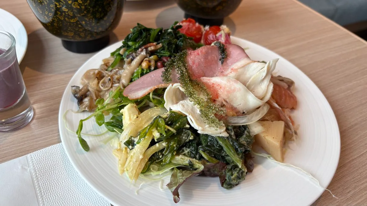 A plate heaped with leaves, sprouts, sea grapes, sautéed mushrooms, cooked spinach, slices of pink cured meat and a baked tomato