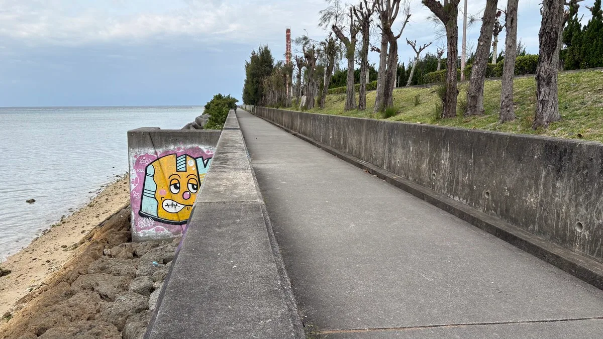 A concrete walkway along a sea wall, with bare trees on a grassy bank to the right and a cartoon face painted on the wall to the left