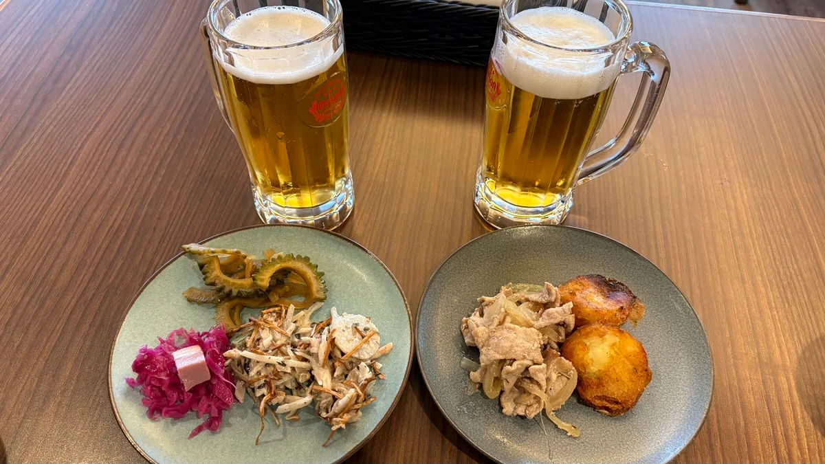 Two mugs of Orion draft beer behind two small plates, one with goya slices, red cabbage and a shredded salad, the other with sliced pork and onion and two browned potatoes