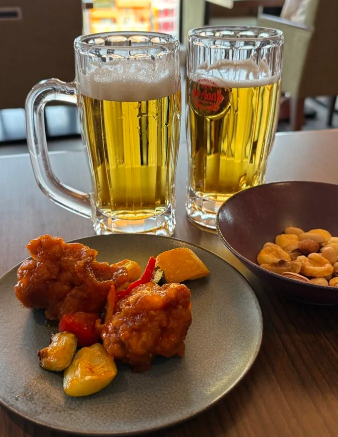 Two mugs of Orion draft beer with a plate of chili-sauce fried chicken and a bowl of mixed nuts