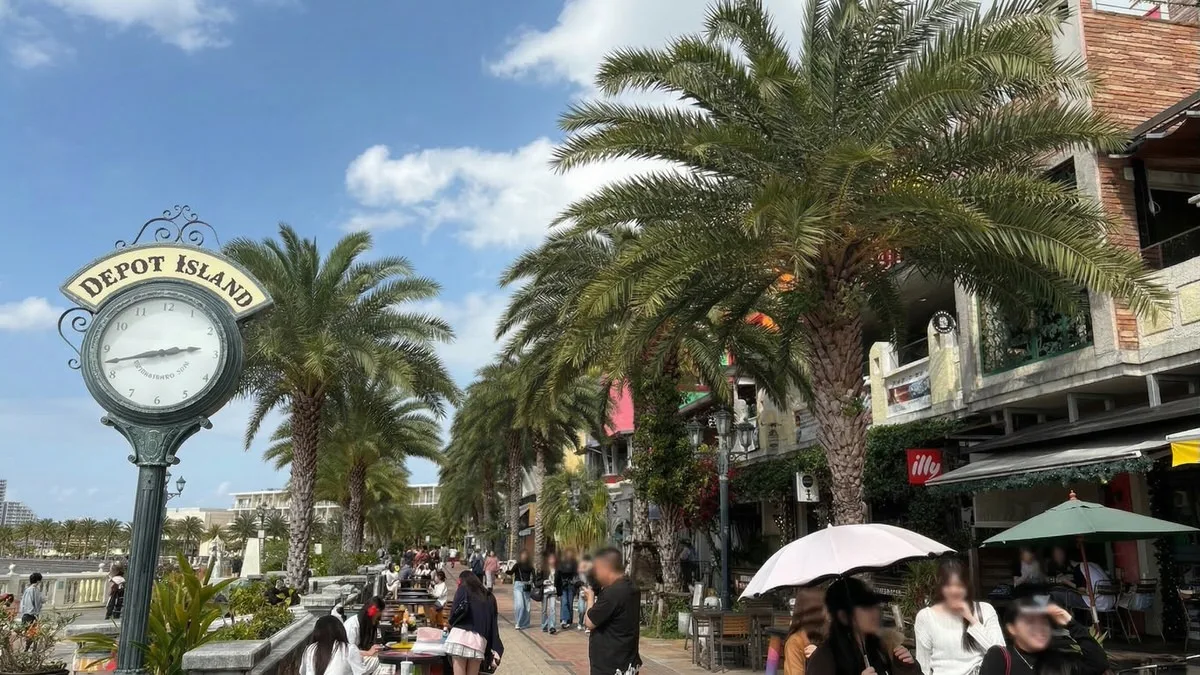 A palm-lined promenade at Depot Island in American Village with an ornamental clock and people walking