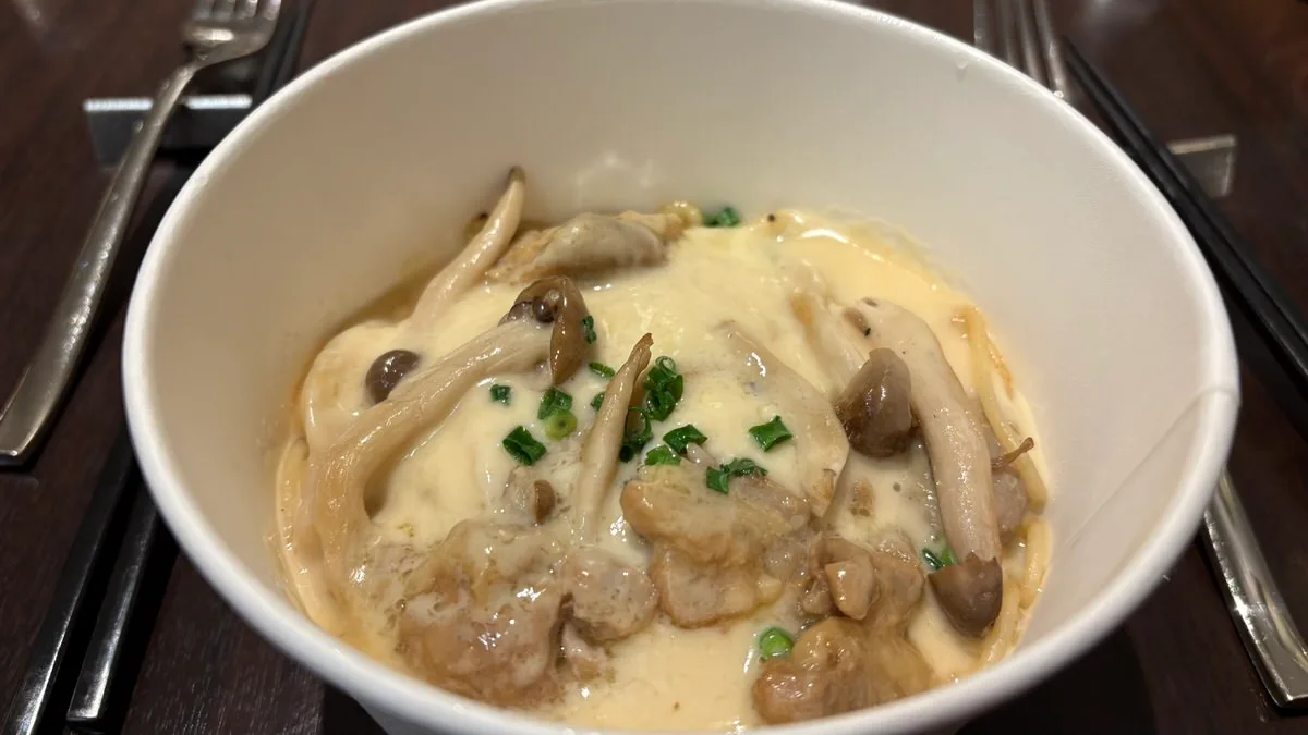 A white takeout bowl of pasta under a cream sauce with chicken pieces, mushrooms and chopped spring onion