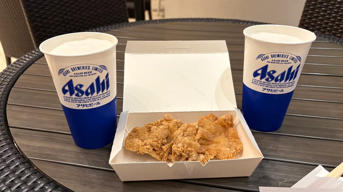 Two paper cups of Asahi draft beer either side of an open white takeout box of fried chicken on a dark table