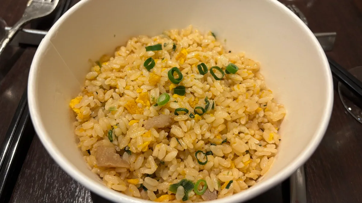 A white takeout bowl of fried rice with egg, spring onion and small pieces of meat