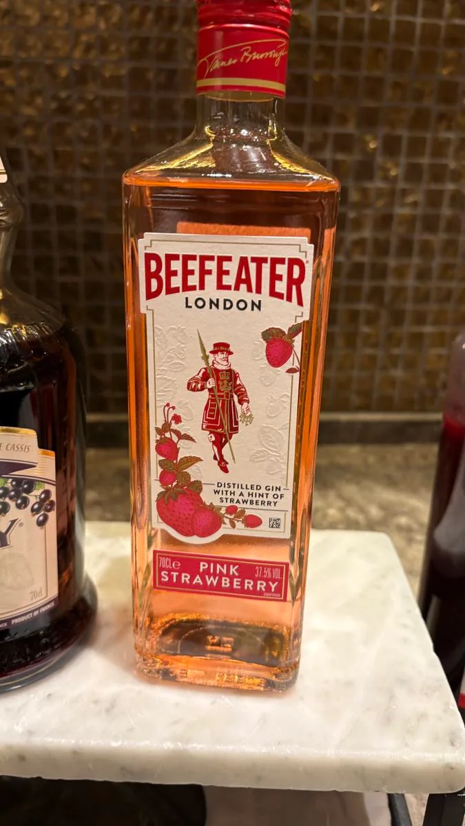 A bottle of Beefeater Pink Strawberry gin standing on a marble counter beside a bottle of creme de cassis