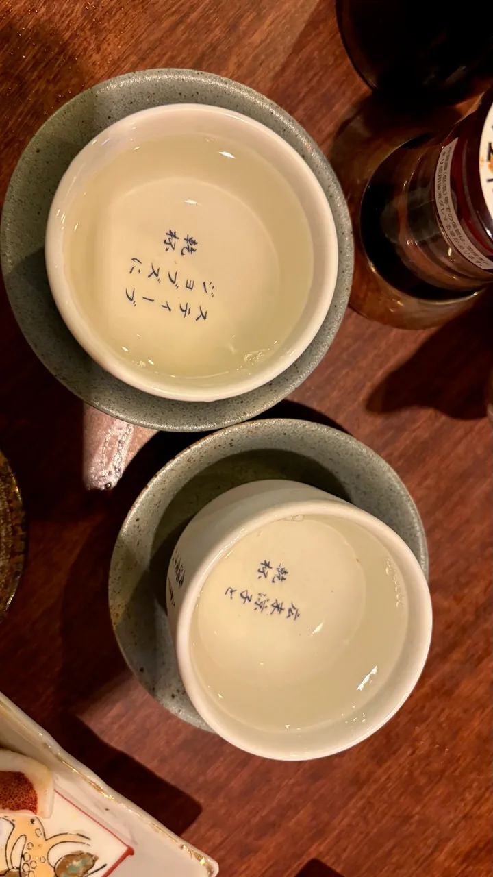 Two white sake cups filled to the brim in speckled ceramic holders on a dark wooden izakaya table