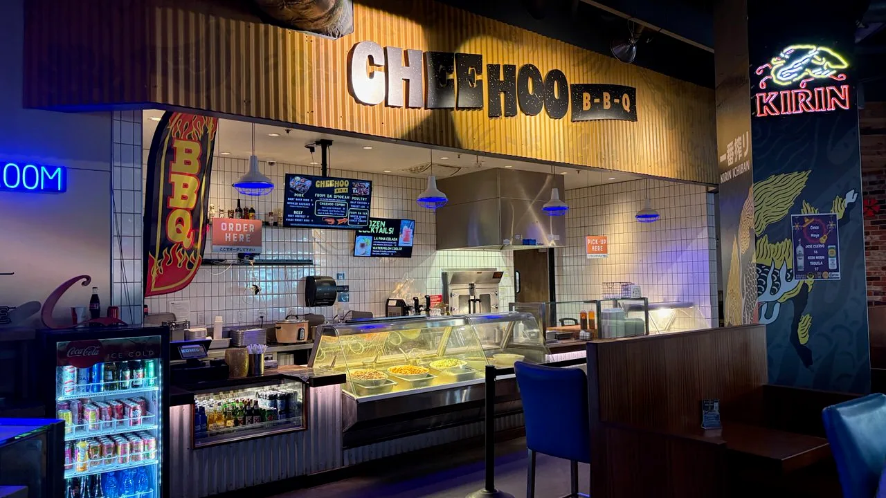 The Chee Hoo BBQ counter under a corrugated-metal sign, with a menu screen, an Order Here sign, a glass serving case and a drinks fridge, and a Kirin Ichiban neon sign on the pillar to the right