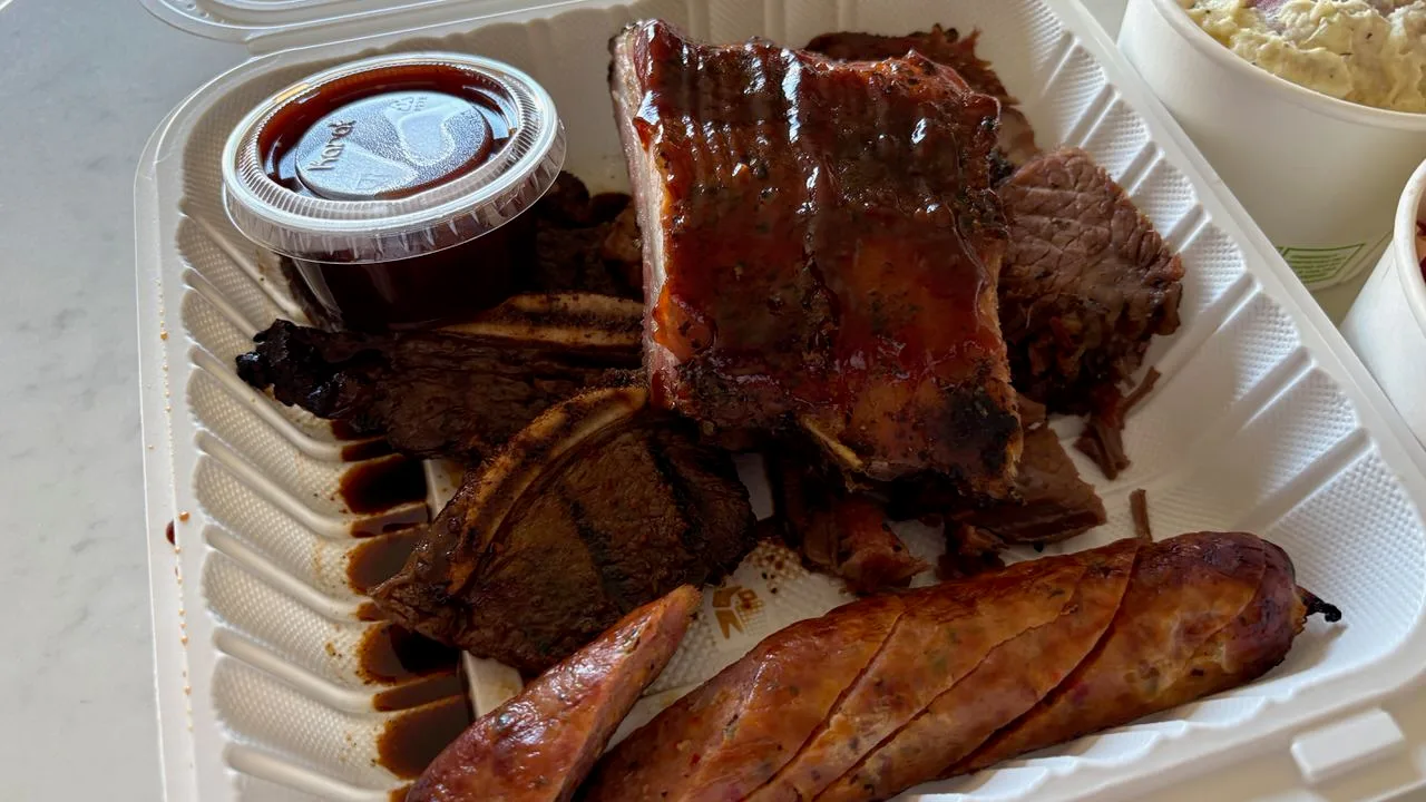 Inside a takeout box: a slab of glazed baby back ribs, bone-in kalbi, slices of brisket and a scored sausage, with a lidded cup of dark sauce