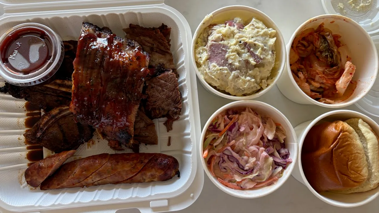 A takeout box of glazed ribs, brisket, kalbi and a scored sausage beside paper cups of potato salad, kimchi, coleslaw and a bread roll