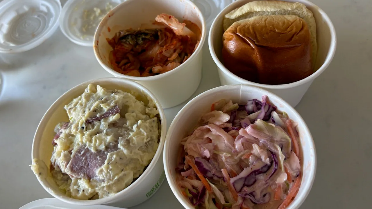 Four paper cups on a white table: kimchi, a bread roll, potato salad, and coleslaw with purple cabbage and carrot