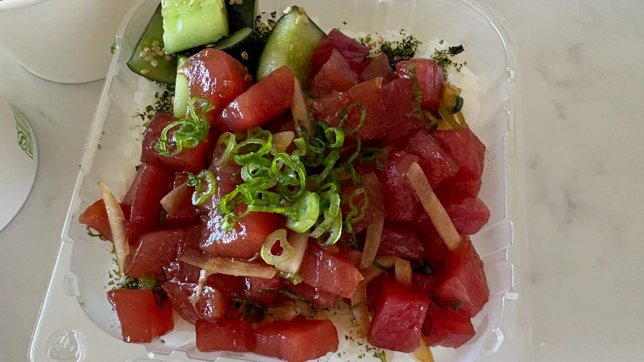 Cubes of ahi in shoyu with sliced green onion and thin strips of onion over rice, and cucumber chunks with sesame at one end, in a clear takeout container