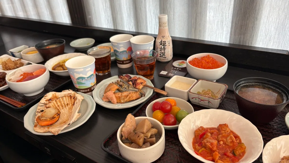 A spread of small buffet dishes on a dark table: a bowl of salmon roe, grilled salmon with a squid skewer, a scallop in its shell, simmered meat and root vegetables, and battered meat in a sweet-and-sour sauce