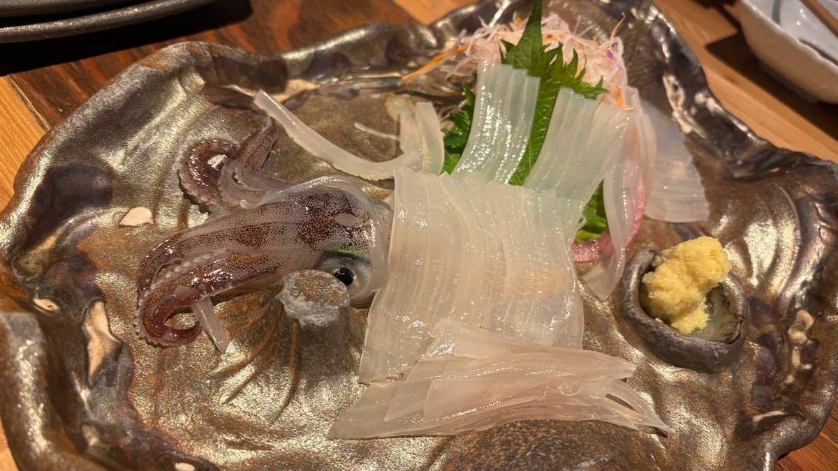 Live squid sashimi sliced translucent on a metal plate, the head and tentacles left whole, with shiso and shredded daikon