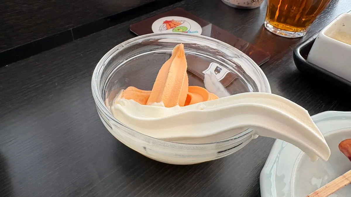 Two soft serve swirls in one glass bowl, orange melon and white milk, on a dark table