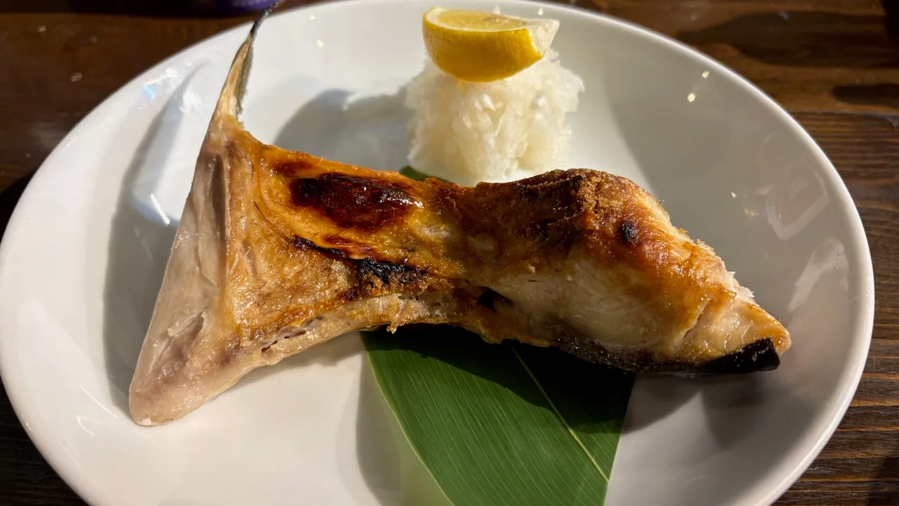 A grilled yellowtail collar on a white plate with grated daikon, a lemon wedge and a green leaf