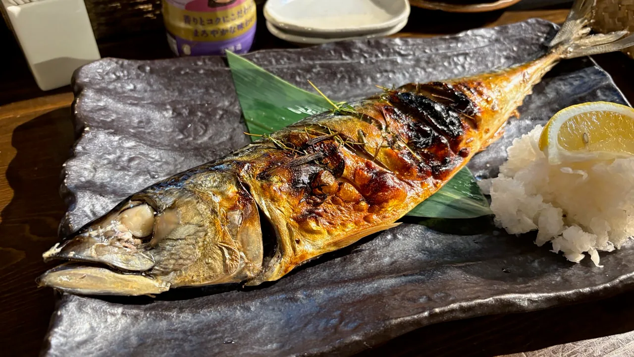 A whole grilled mackerel head to tail on a dark slate plate with grated daikon and a lemon wedge