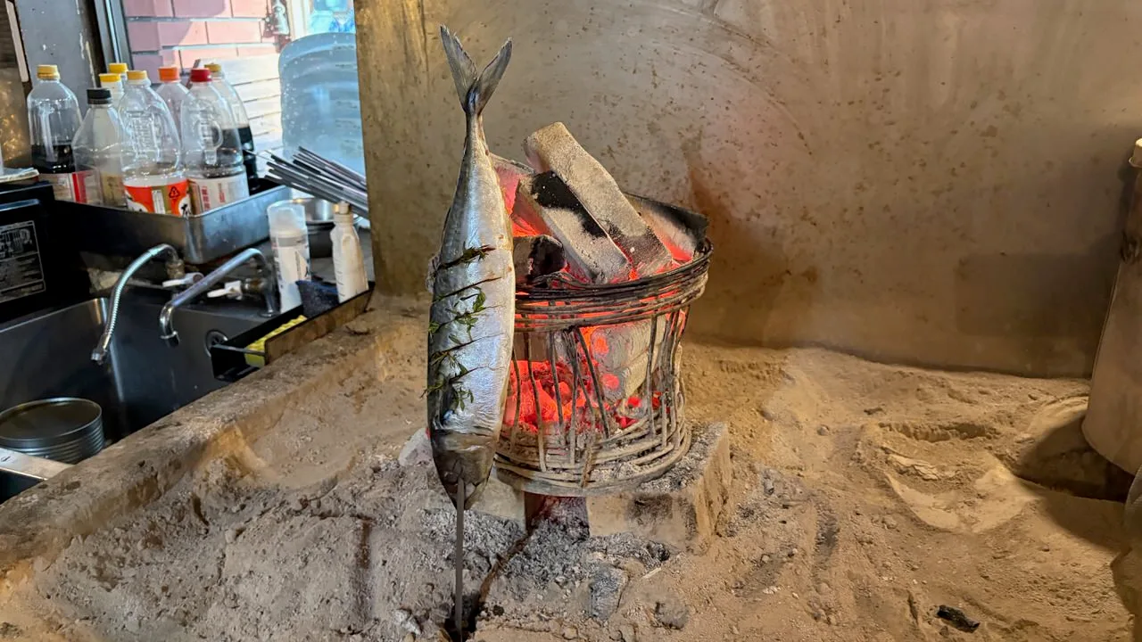 A whole mackerel on a long metal skewer standing upright in sand next to a wire basket of glowing charcoal