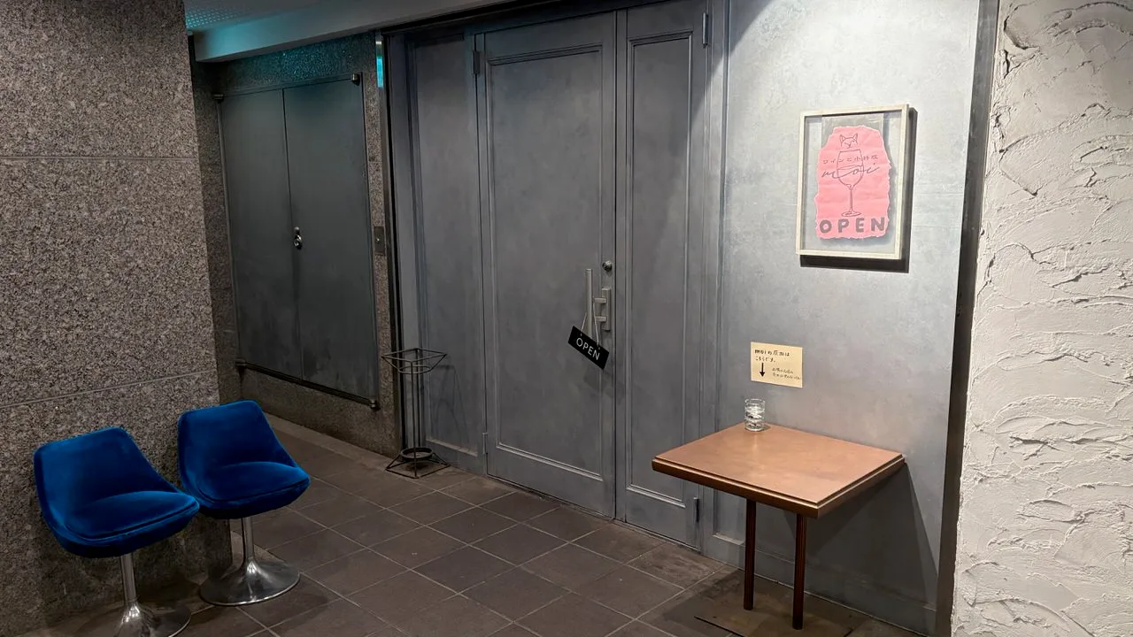 A grey door on a grey landing with a small black OPEN tag on the handle, a framed pink sign on the wall and two blue velvet chairs