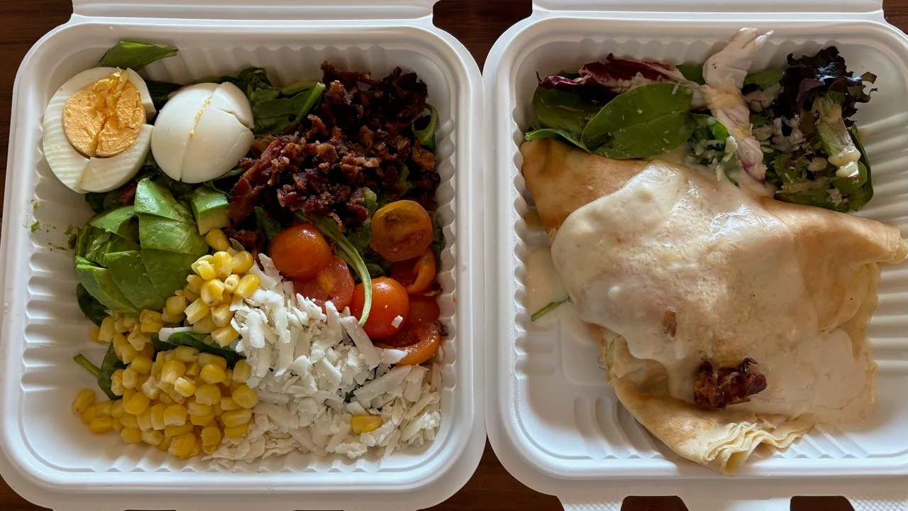 Two open takeout boxes on a wooden table: a Cobb salad of spinach, boiled egg, bacon bits, avocado, cherry tomatoes, corn and crumbled white cheese, and a folded crepe under white sauce with mixed leaves