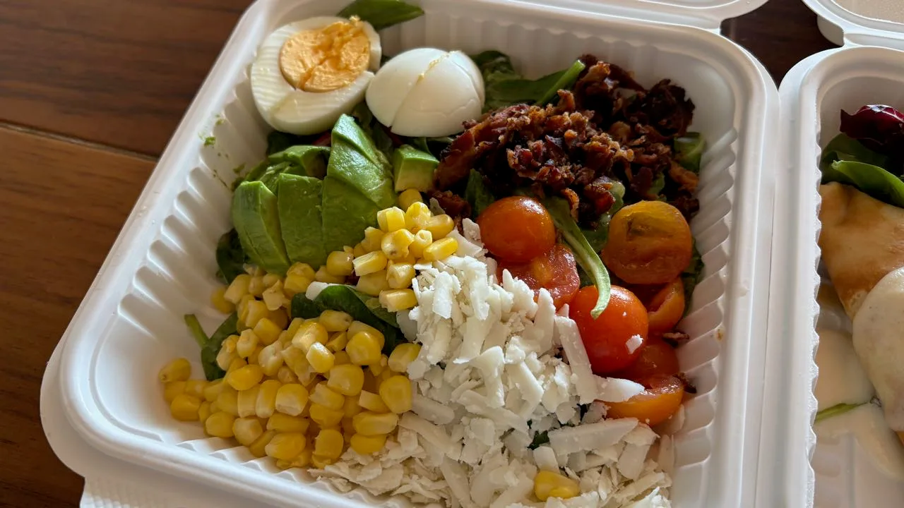 Close-up of the Cobb salad: boiled egg halves, crisp bacon pieces, avocado slices, red and yellow cherry tomatoes, corn and crumbled white cheese over spinach