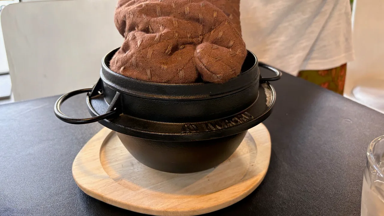 A black lidded pot with ring handles on a round wooden board, a hand in a quilted brown oven mitt pressing down on the lid