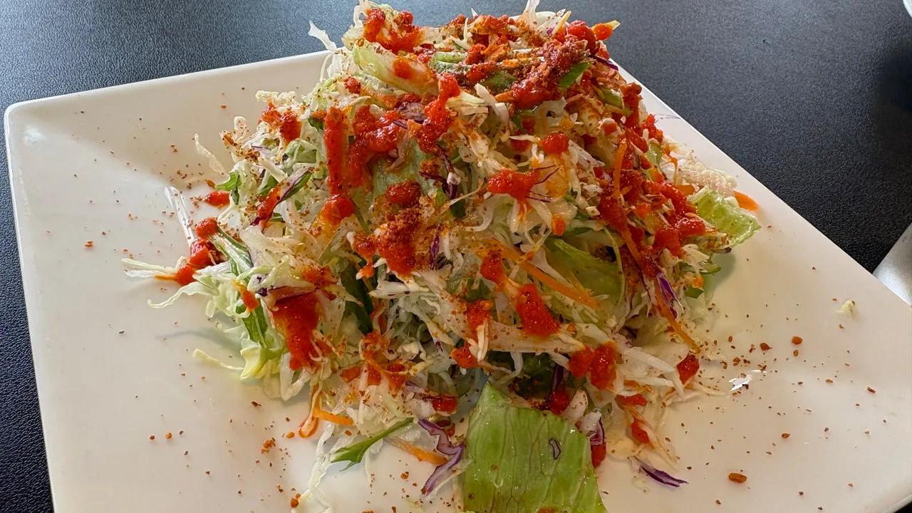 A mound of shredded cabbage, lettuce, red cabbage and carrot on a square white plate, drizzled with orange-red dressing and dusted with red spice