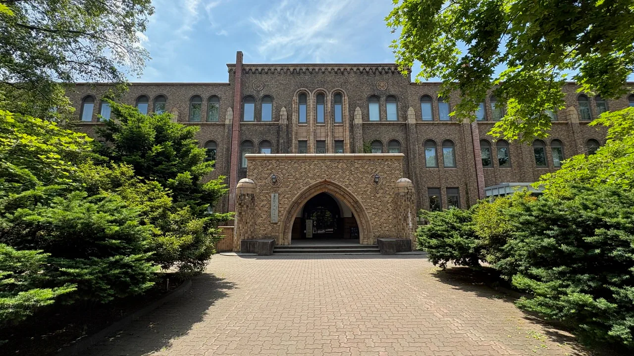 A brown brick university building with a pointed stone archway entrance, framed by clipped hedges and a brick path