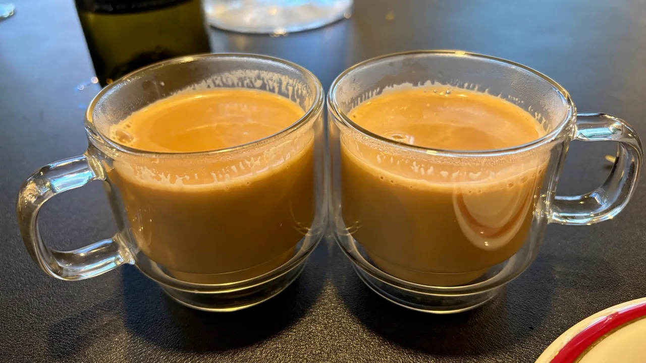 Two clear glass mugs of milky chai side by side on a dark table
