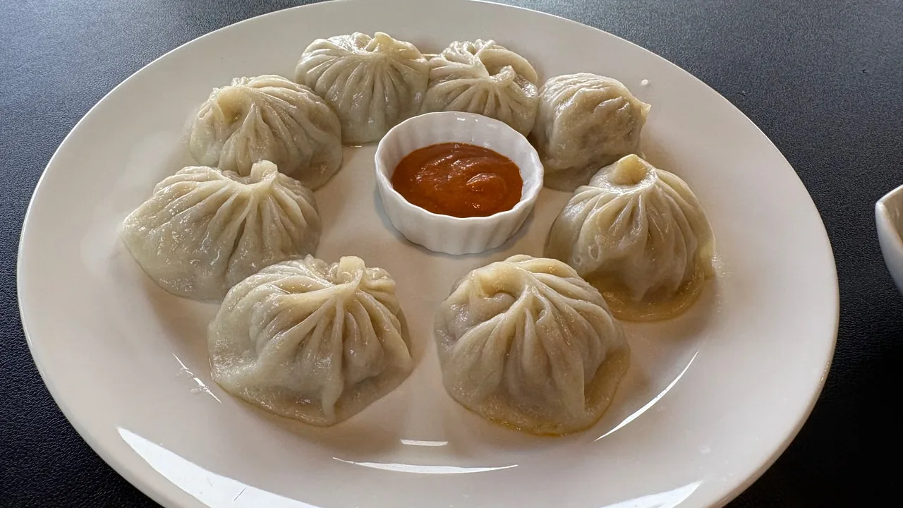 Eight pleated steamed dumplings arranged in a ring on a white plate around a small fluted dish of orange-red sauce