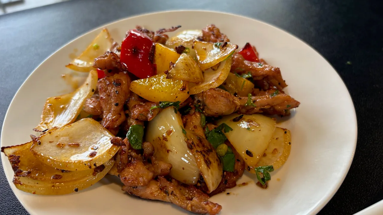 Stir-fried chicken neck meat with wedges of onion and pieces of red and yellow pepper, scattered with chopped herbs, on a white plate