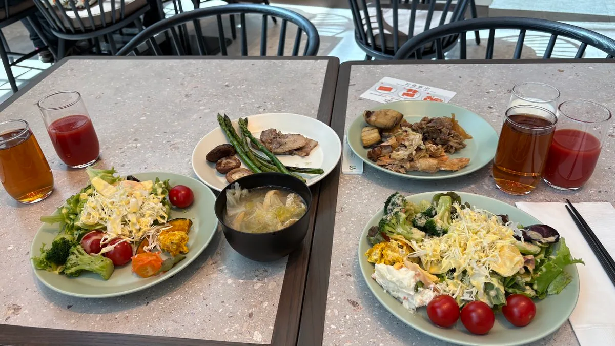 Two breakfast settings on a stone-topped table: salad plates with broccoli and cherry tomatoes, a plate of grilled asparagus with mushrooms, a black bowl of miso soup, and glasses of tea and tomato juice