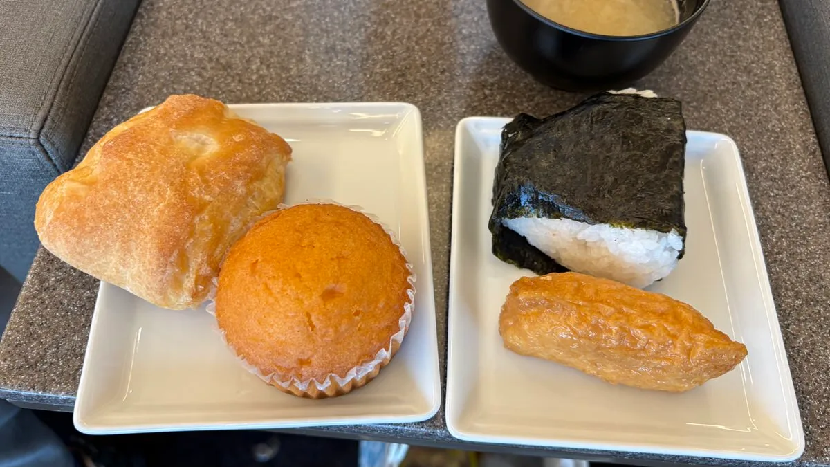 Two white square plates on a stone-topped side table: a pale puffy bread roll and a golden muffin in a paper case on one, a nori-wrapped rice ball and an inari sushi on the other