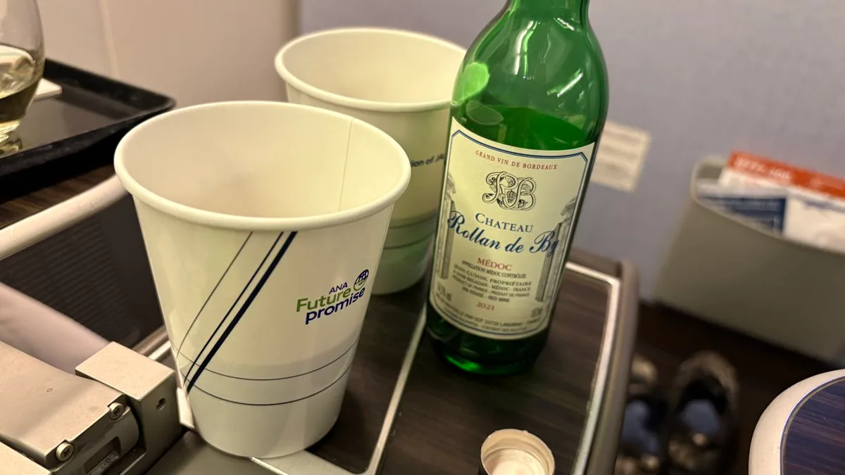 A green bottle of red wine standing on an aircraft tray table next to two white paper cups printed with an airline logo