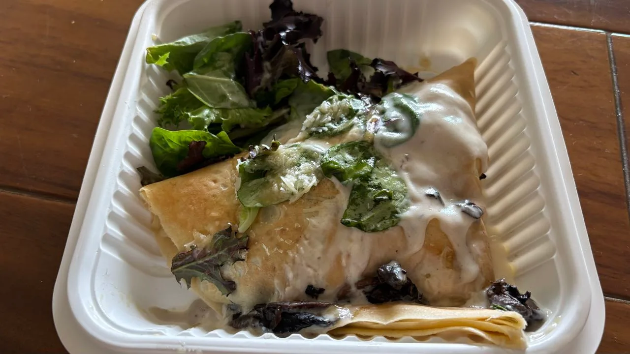 A folded crepe under cream sauce with grated cheese and greens in a white takeout box, with a side salad