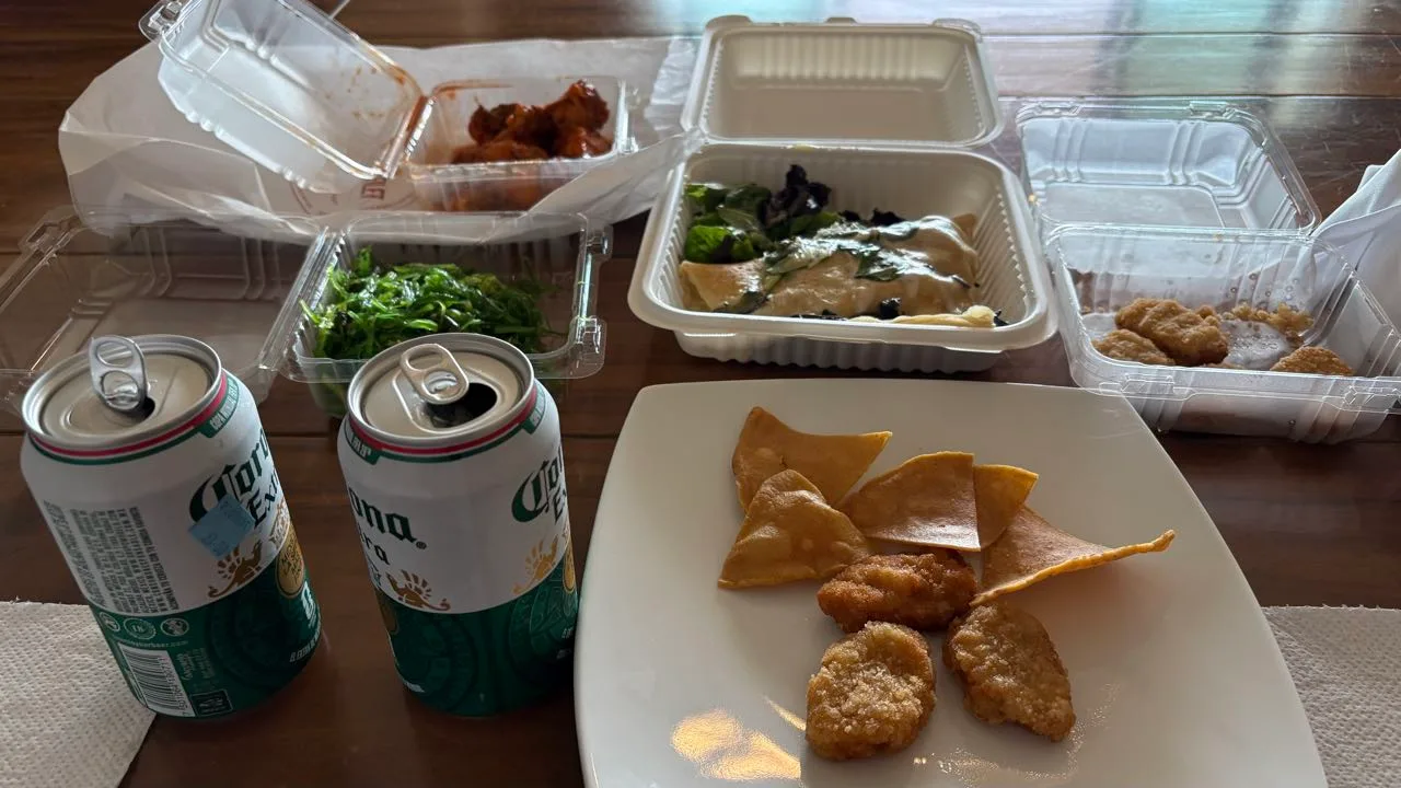 Two open Corona cans and takeout boxes of wings, seaweed salad and nuggets on a wooden table, with tortilla chips and nuggets on a white plate
