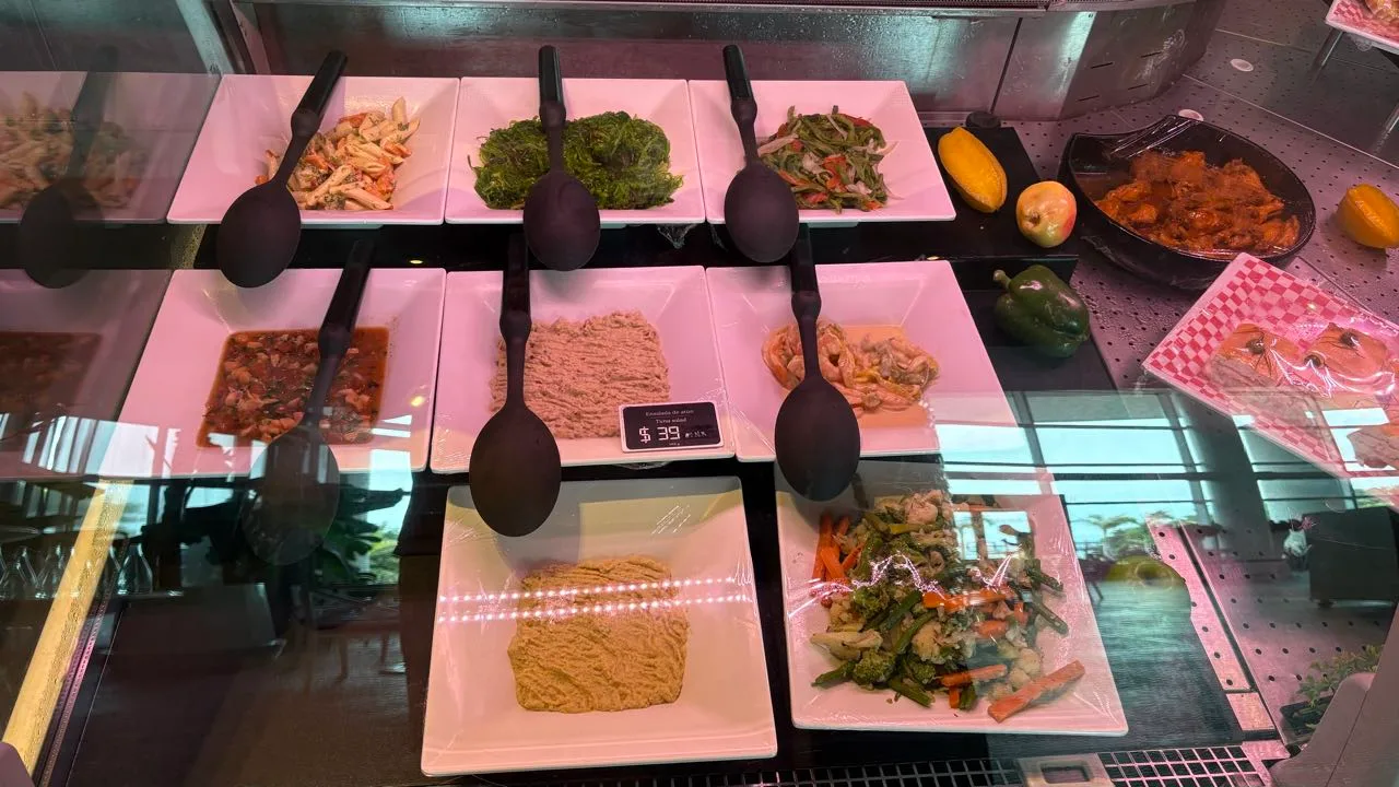 Square white plates of prepared salads behind glass at a deli counter, each plate with a black serving spoon