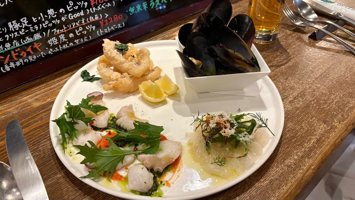 A white plate with sliced raw fish under greens and green sauce, thin slices topped with dill, and golden fried squid with two lemon wedges, next to a square bowl of mussels