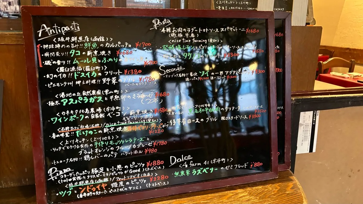 A large blackboard menu handwritten in coloured chalk in Japanese, with sections for antipasti, pasta, secondo, pizza and dolce