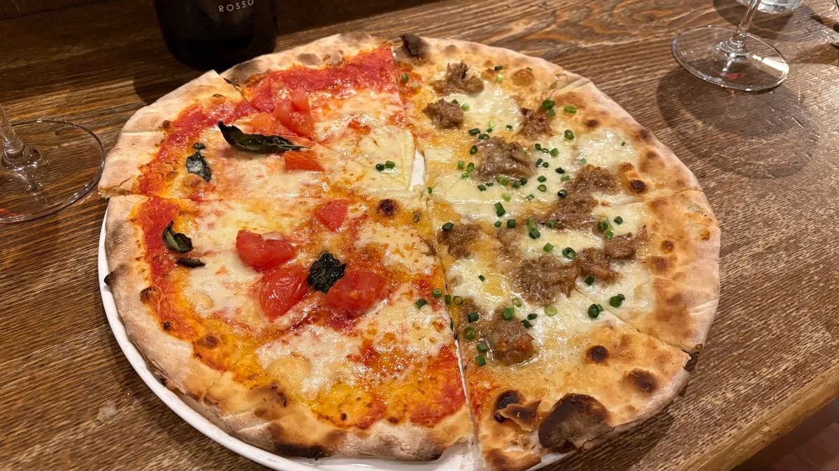 A whole pizza on a white plate split down the middle, tomato and basil on one side, white cheese base with braised pork and green onion on the other, a red wine bottle behind