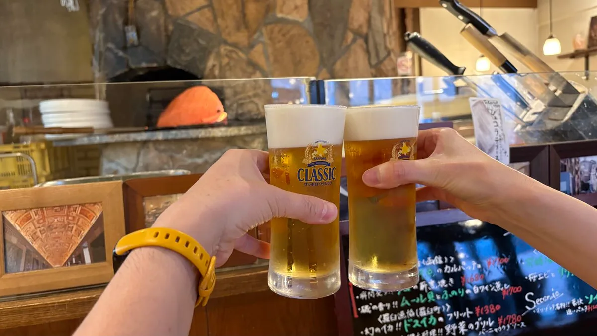 Two tall glasses of pale lager being clinked together over a counter, with the mouth of a brick oven behind glass in the background