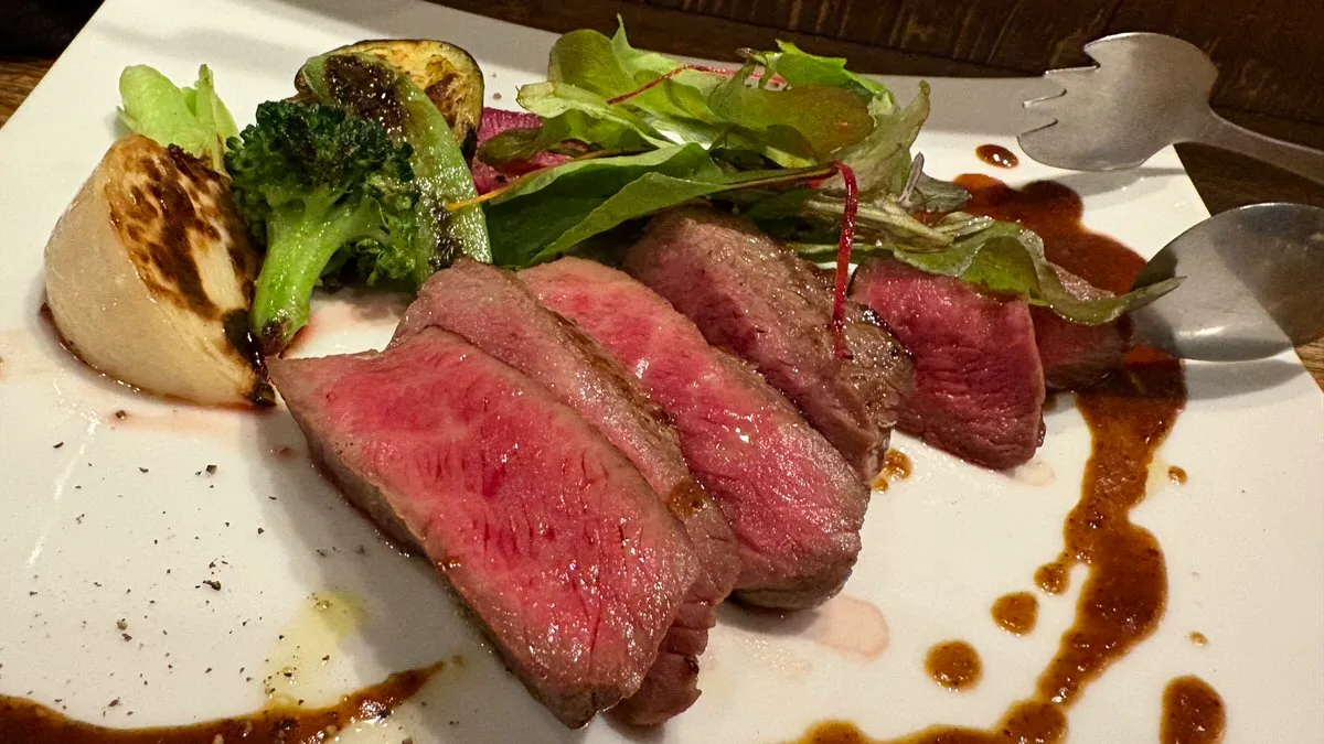 Five thick slices of beef, pink in the middle, on a white plate with grilled onion, broccoli, courgette, salad leaves and a dark sauce
