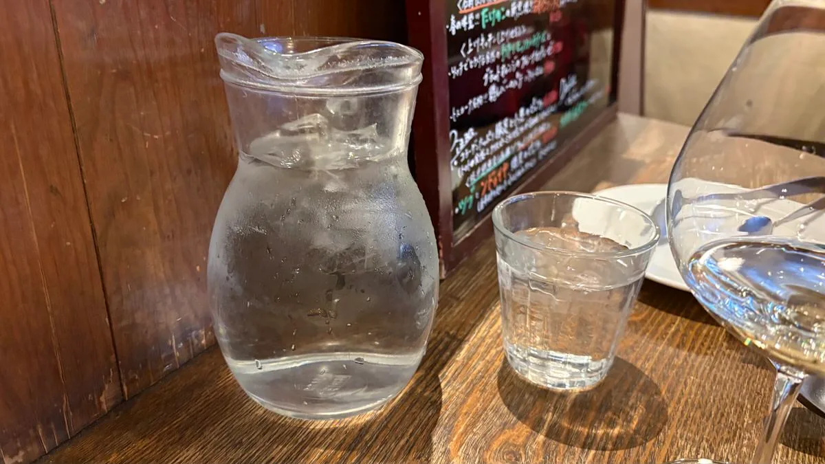 A small glass pitcher of iced water beside a tumbler and a wine glass on a wooden counter