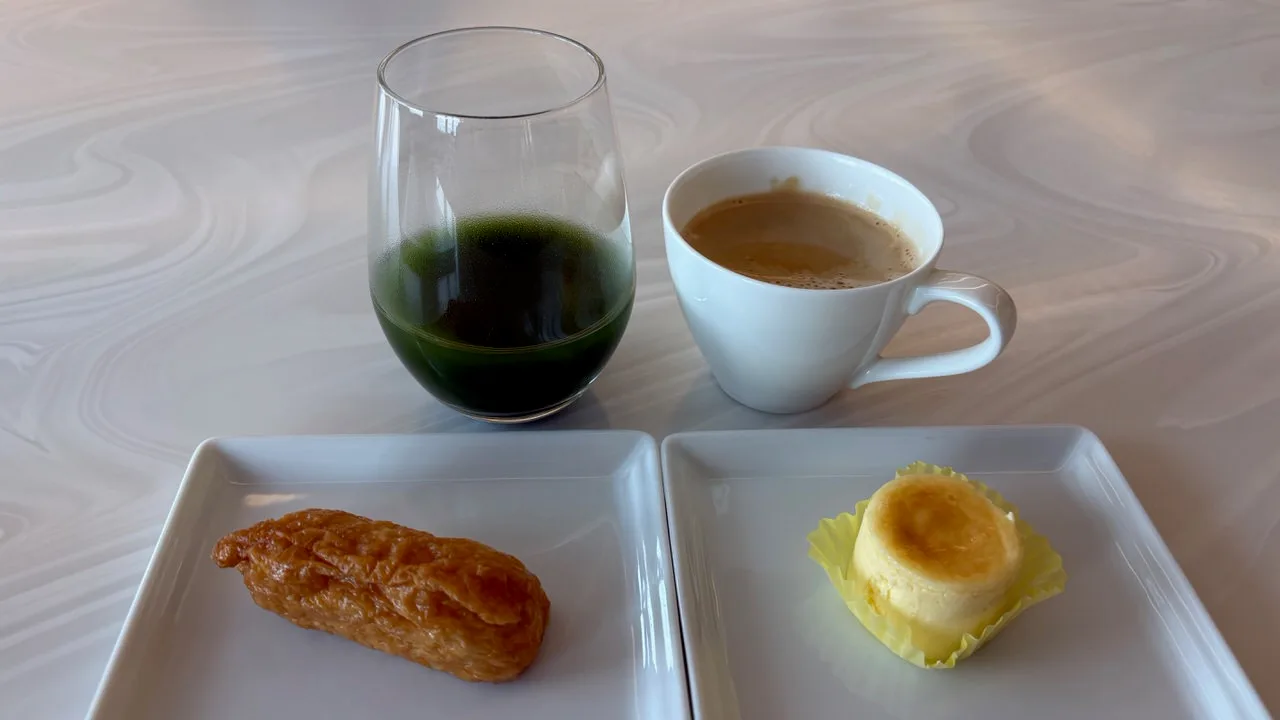 A glass of dark green kale juice, a white cup of coffee, an inari sushi on one square plate and a small cheese souffle in a yellow paper case on another