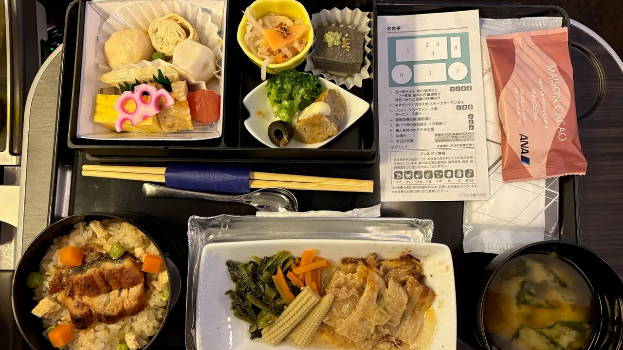 An airline meal tray from above with a boxed assortment of cold dishes, a hot plate of sliced pork with greens and baby corn, a bowl of rice with eel, miso soup, chopsticks and a menu card