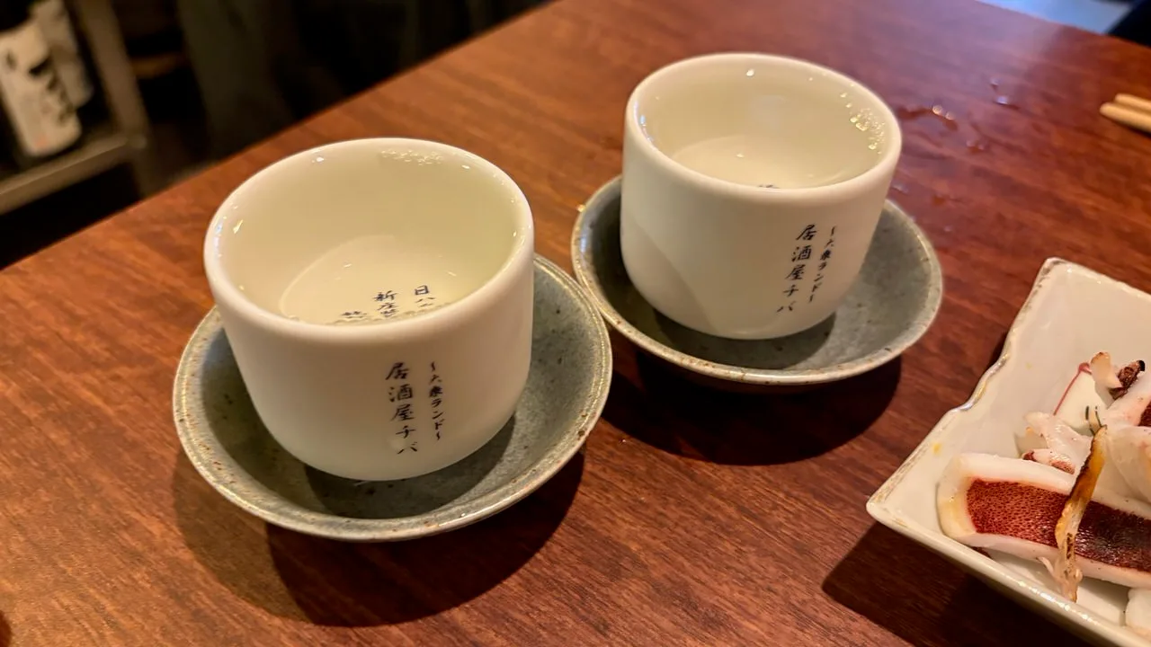 Two white sake cups filled to the brim on speckled grey saucers on a dark wooden table, with a dish of grilled squid beside them