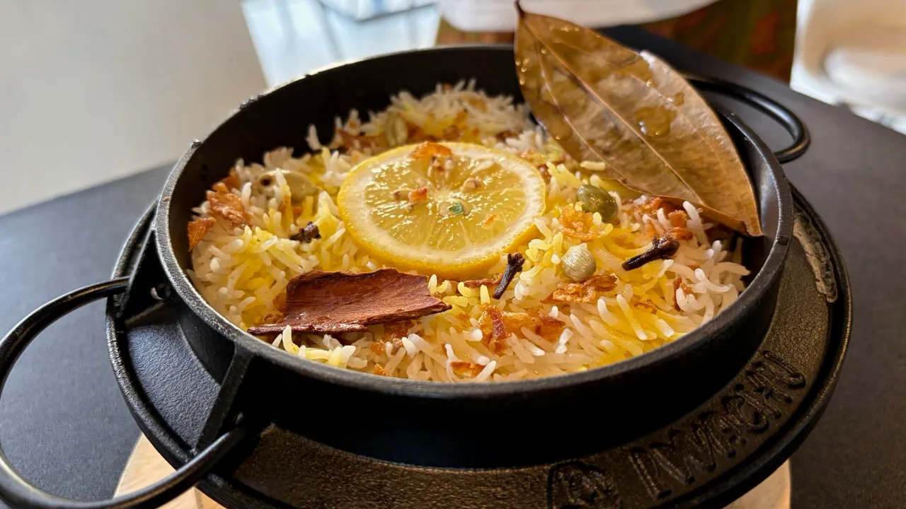 A black clay pot of basmati biryani topped with a lemon slice, two bay leaves, a piece of cinnamon bark, cloves and fried onion
