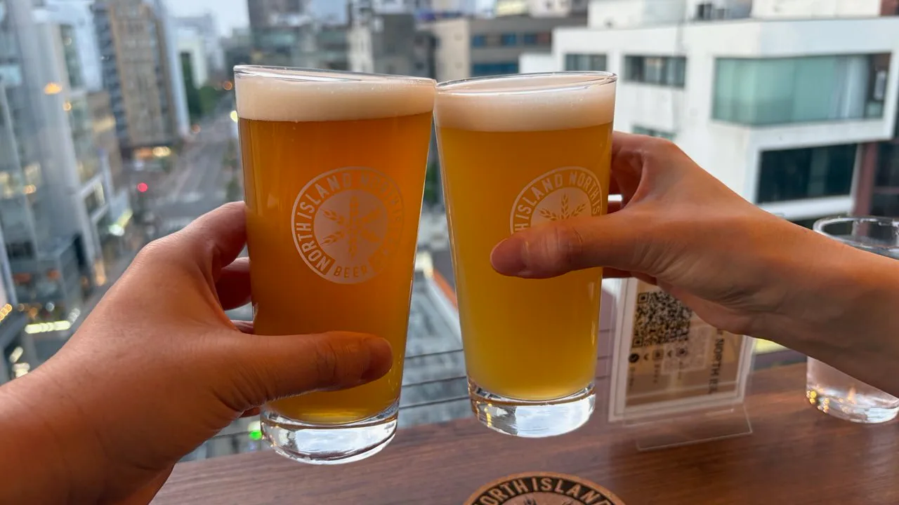 Two glasses of hazy pale beer held up against a window with city buildings ten floors below at dusk