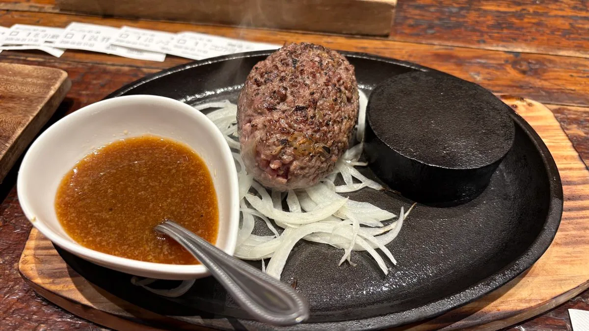 A tall beef patty on a black iron plate over sliced raw onion, with a hot stone and a bowl of brown sauce