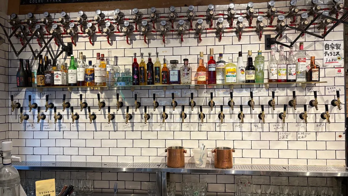 A white tiled wall covered in rows of beer taps with handwritten labels, spirit bottles on a shelf above
